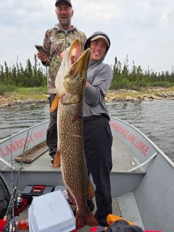 Two people on a boat hold a large fish, with fishing gear and trees visible in the background.