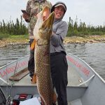 Two people on a boat hold a large fish, with fishing gear and trees visible in the background.