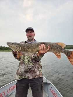 A man standing in a boat holds a large fish with both hands on a lake, with trees visible in the background.