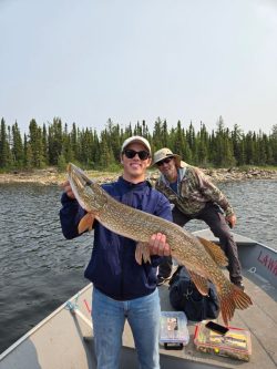 Man standing on a boat holding a large fish, with another man smiling in the background; lake and trees visible.