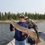Man standing on a boat holding a large fish, with another man smiling in the background; lake and trees visible.