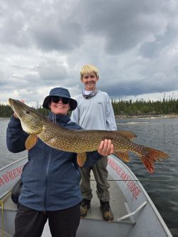 Person in sunglasses and jacket holding a large fish on a boat with another person standing behind, lake and trees in background.