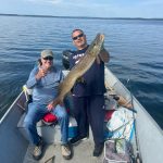 Two men in a boat on a lake; one is seated, giving a thumbs up, while the other holds a large fish.