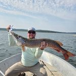 Man in a boat holding a large fish with both hands on a lake under a partly cloudy sky.