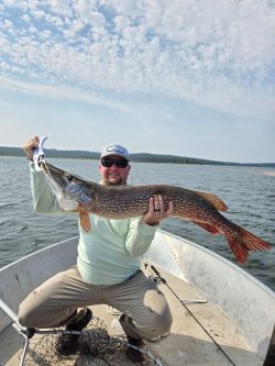 Man in a boat holding a large fish with both hands on a lake under a partly cloudy sky.