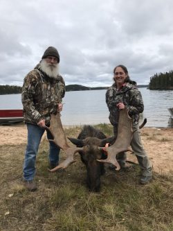 Two people in camo clothing pose by a large moose with antlers on a lakeshore, with trees and a boat in the background.