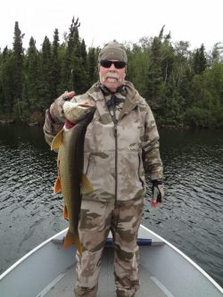 Man in camouflage clothing stands on a boat holding a large fish, with trees and water in the background.