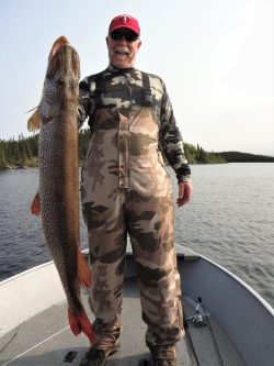 Man in camouflage gear standing on a boat holding a large fish beside a body of water with forested shoreline.
