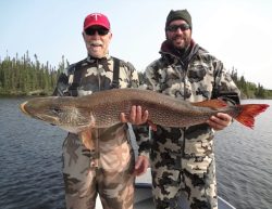 Two men in camouflage outfits hold a large fish on a boat, with a lake and trees in the background.