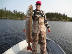Man in camouflage outfit stands on a boat holding a large fish, with a lake and trees in the background.