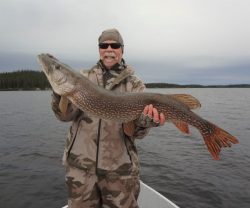 Man wearing camouflage clothing holds a large fish while standing on a boat on a lake with forested shore in background.