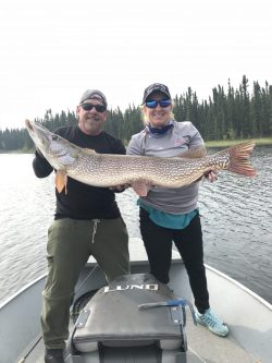 Two people on a boat hold a large fish together, with trees and water visible in the background.