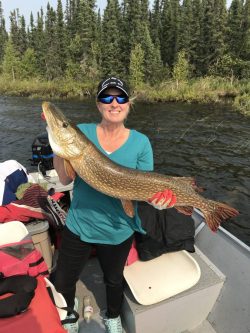 A woman on a boat holds a large fish with both hands, smiling at the camera. Trees and water are in the background.