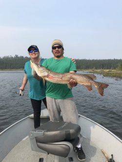 Two people standing on a boat hold a large fish together on a lake, with trees visible in the background.