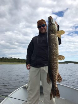 Person standing on a boat holds a large fish upright with one hand on a lake under a cloudy sky.