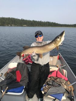 Person standing in a boat on a lake, holding a large fish with both hands, surrounded by fishing gear.