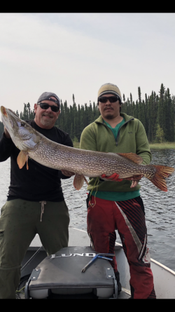 Two people on a boat hold a large fish together, with trees and water in the background.
