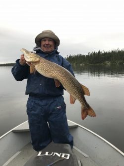 Person in rain gear holding a large fish on a boat in a calm lake with trees in the background.