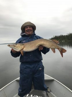 Person in a rain jacket holds a large fish on a boat in a calm lake, with trees visible in the background.