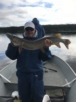 Person in blue outerwear holding a large fish on a boat, with another person facing away in the background on a calm lake.