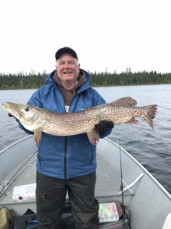 A man standing in a boat holds a large fish horizontally in front of him on a lake.