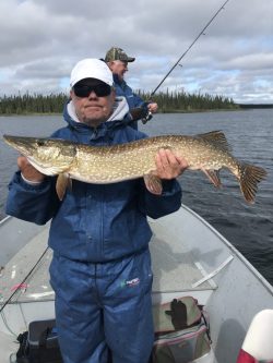 Person in blue rain gear holding a large fish on a boat, with another person fishing in the background on a lake.