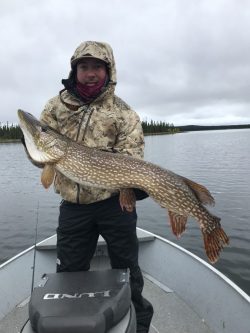 Person in camouflage jacket holding a large northern pike fish on a boat in a lake under an overcast sky.