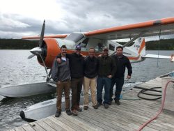 Five men stand on a dock next to an orange and white floatplane on a lake under cloudy skies.