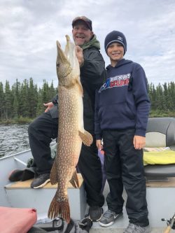 Two people pose on a boat; one holds up a large Northern pike fish, with trees and water in the background.