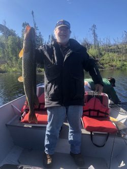 A man standing on a boat holds a large fish upright; another person is in the background near the water.
