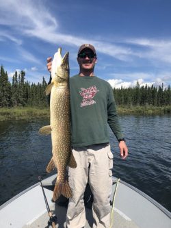 Person standing on a boat holding a large fish with trees and water in the background.