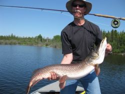 Man holding a large fish beside a lake, with fishing rod balanced on his shoulders, trees in background.