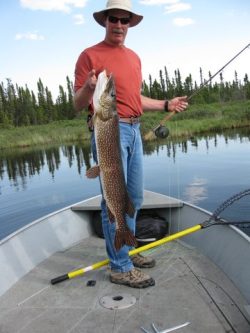 Man standing in a boat holding a large fish, with fishing gear and a net on the boat floor.