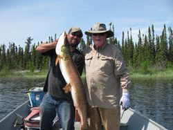 Two men in a boat holding a large fish, with trees and water in the background on a sunny day.