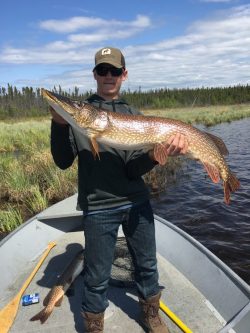 Person standing on a boat holds a large northern pike fish; another fish and fishing gear are on the boat floor.