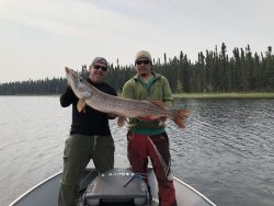 Two men standing on a boat holding a large fish, with a forested shoreline in the background.