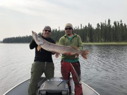 Two men standing in a boat on a lake, holding a large fish together with trees in the background.