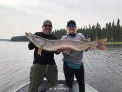 Two people standing on a boat hold a large fish together, with water and trees visible in the background.