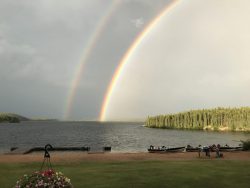 Two rainbows arc over a lake with boats docked by the shore and a flower basket in the foreground.