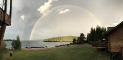A rainbow arches over a lakeside landscape with trees, a dock, and cabins under a partly cloudy sky.