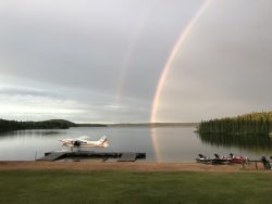 A floatplane and boats are docked on a lake with a double rainbow in the cloudy sky.