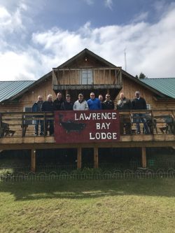 A group of people stand on the deck of Lawrence Bay Lodge in front of a large sign on a sunny day.