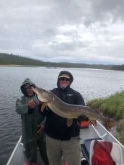 Two people in rain gear on a boat hold a large fish over a lake with a forested shoreline under cloudy skies.