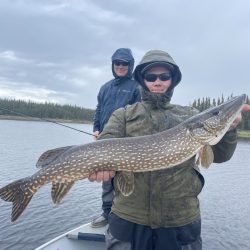 Two people on a boat in rainy weather; one holds a large northern pike fish while the other stands behind.