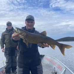 Person holding a large fish on a boat with another person standing behind; lake and cloudy sky in the background.