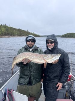 Two men in raincoats on a boat holding a large fish, with a lake and forested shoreline in the background.