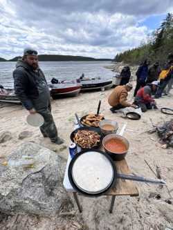 People prepare and cook food on the sandy shore of a lake, with boats docked nearby and cloudy skies overhead.