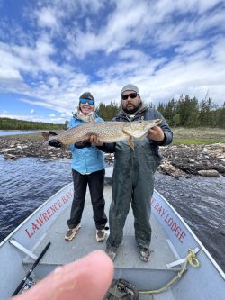Two people on a boat holding a large fish, with trees and water in the background under a partly cloudy sky.