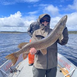 Person in a hooded jacket holds a large fish on a boat, with water and cloudy sky in the background.