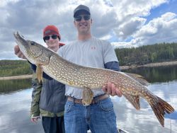 Two people on a boat hold a large northern pike fish with a forested shoreline and cloudy sky in the background.